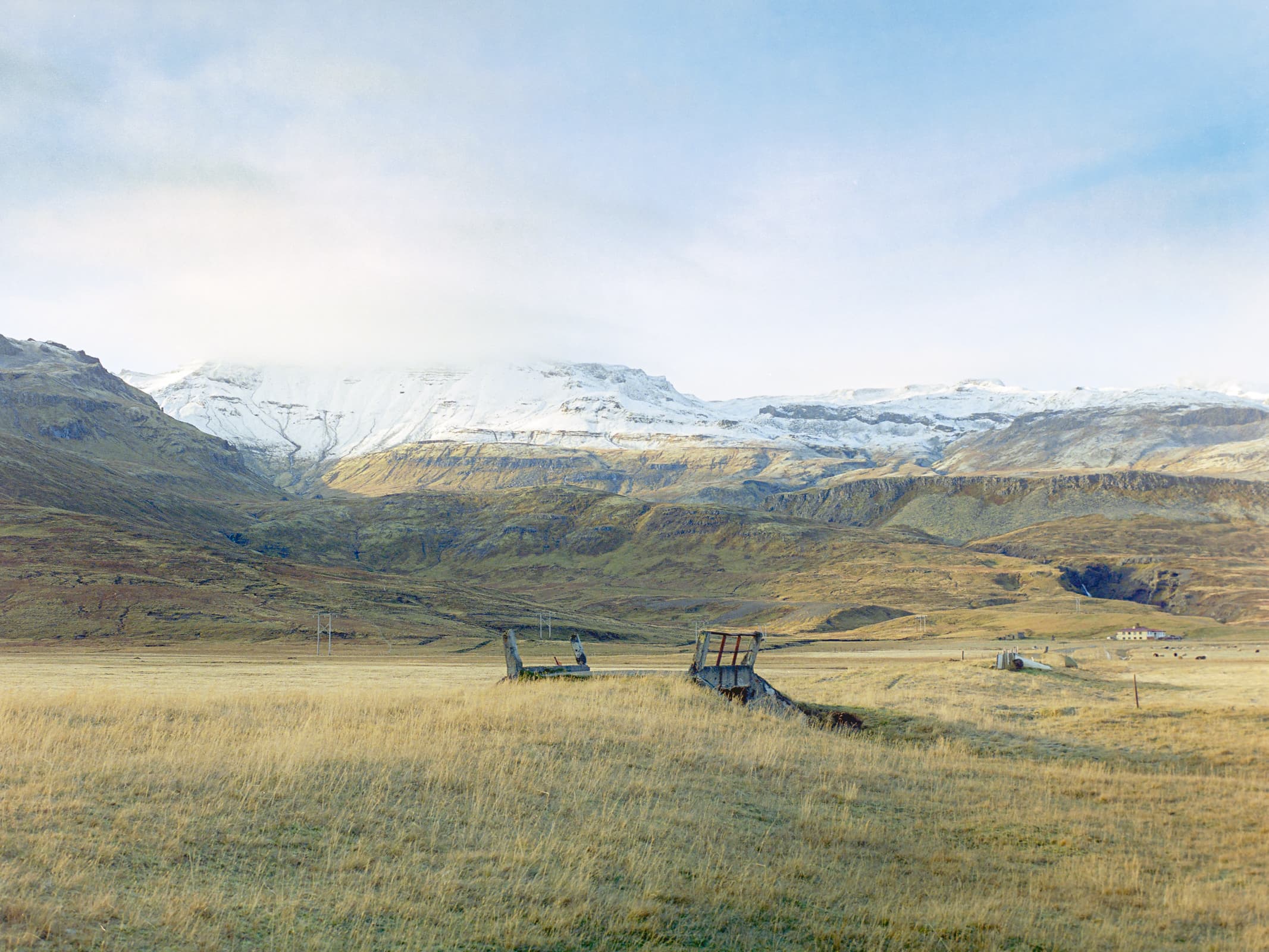 a small, old bridge which is no longer in use sits in a lush green valley with a backdrop of dramatic, snowcapped peaks.