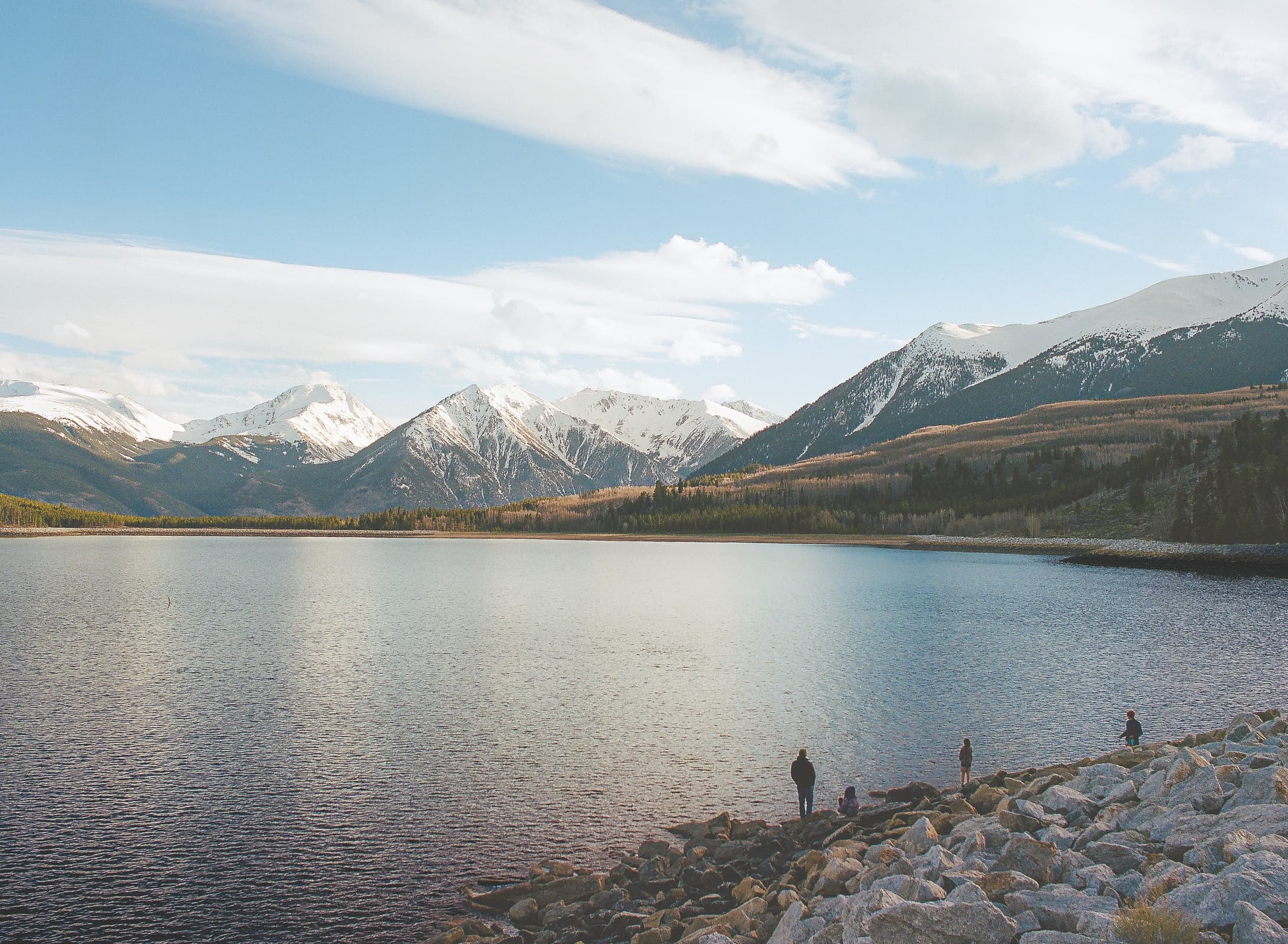 4 people standing on the rocky shore of a high-alpine reservoir surrounded by snowcapped peaks