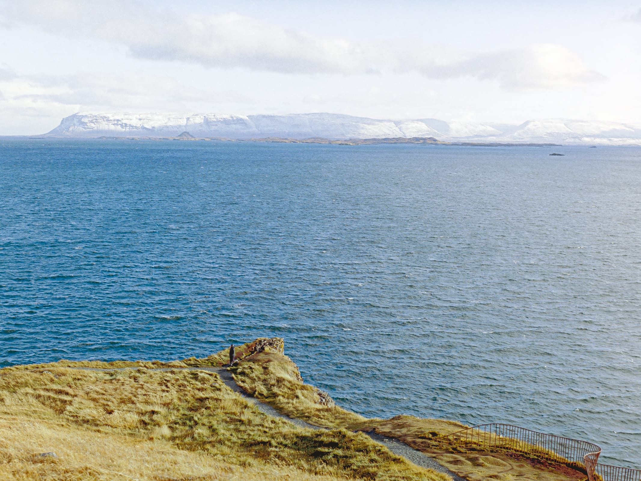 a person standing at a dramatic point on a grassy hillside that juts out over the sea. across the sea are snow-covered mountains. the sea is peppered with islands of varying sizes.